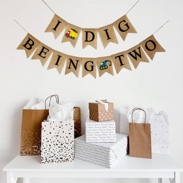 This two tiered banner spells out the words I Dig Being Two. Between the words I and Dig there is a red and yellow dump truck image. The second line reads Being Two with a blue digger truck in between the words Being and Two.