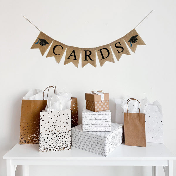 A burlap banner made from 4.5 by 6.5 inch fishtail shaped flags. Each flag has a pocket across the top for stringing. This banner says CARDS in black capital letters. There is a hand stenciled image of a black graduation cap with a blue tassel on each end of the name. It is strung on a jute string that is 110 inches in length.