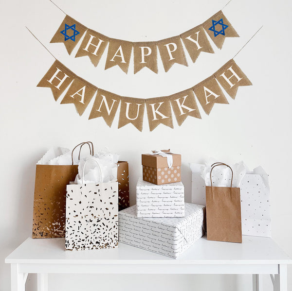 A burlap banner made from 4.5 by 6.5 inch fishtail shaped flags. Each flag has a pocket across the top for stringing. The top line of this banner says HAPPY in white capital letters and has a hand stenciled image of a blue Star of David on each end of the word. The bottom line says HANUKKAH in white capital letters. It is strung on two 110 inch jute strings.
