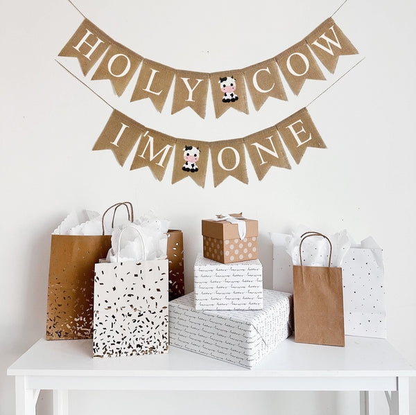 A burlap banner made from 4.5 by 6.5 inch fishtail shaped flags. Each flag has a pocket across the top for stringing. The top line says HOLY COW in white capital letters and has a hand stenciled image of a black and white sitting baby cow between the words. The bottom line says I&#39;M ONE and has an image of the baby cow between the words. It is strung on two 110 inch jute strings.