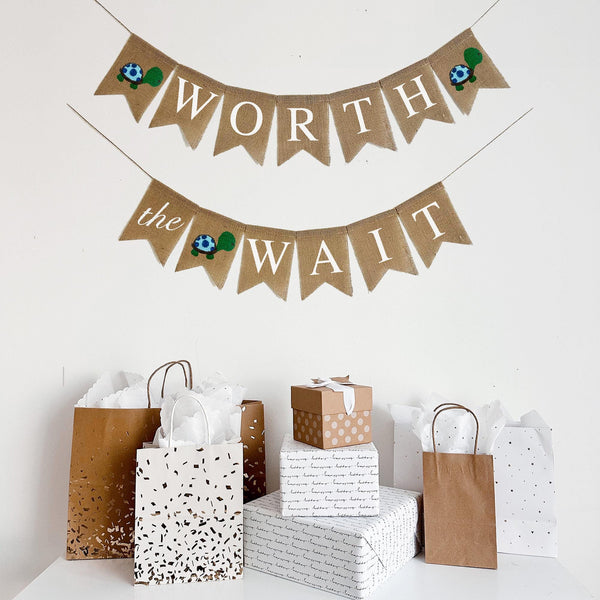 This &quot;Worth the Wait&quot; two tiered burlap banner is perfect for a baby shower. On the first flag there is a blue and green hand stenciled turtle. Following the turtle image the banner reads WORTH with another turtle on the end. The second line starts with a flag reading &quot;the&quot; in script font, followed by another hand stenciled turtle flag and ends with the flags read WAIT.