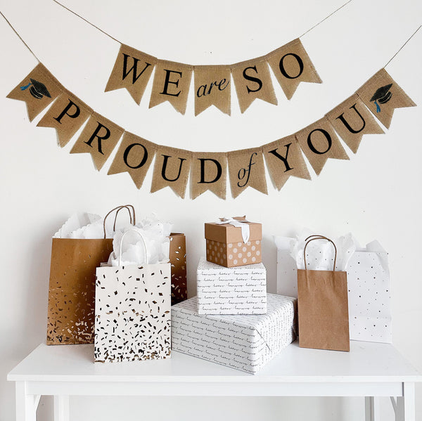 A burlap banner made from 4.5 by 6.5 inch fishtail shaped flags. Each flag has a pocket across the top for stringing. The top line says WE ARE SO in black capital letters with the word ARE on one flag. The bottom line says PROUD of YOU in black capital letters with a hand stenciled image of a graduation cap on each end. Each line is strung on a jute string that is 110 inches in length.