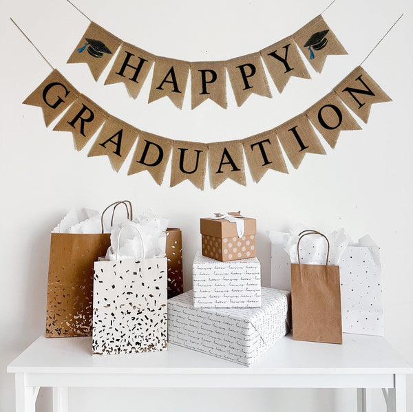 A burlap banner made from 4.5 by 6.5 inch fishtail shaped flags. The top line says HAPPY in black capital letters and has a hand stenciled image of a black graduation cap with a blue tassel on each end. The bottom line says GRADUATION in black capital letters. Each line is strung on a jute string that is 110 inches in length.
