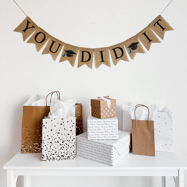A burlap banner made from 4.5 by 6.5 inch fishtail shaped flags. Each flag has a pocket across the top for stringing. It says YOU DID IT in black capital letters with a hand stenciled graduation cap with a blue tassel in between each word. It is is strung on a jute string that is 110 inches in length.