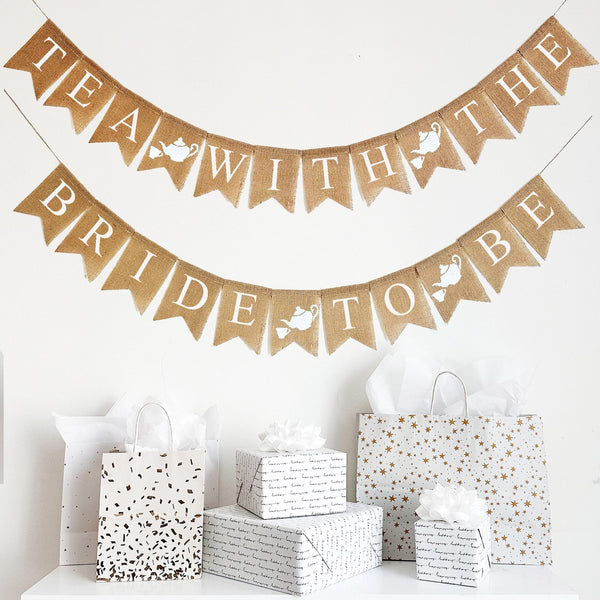This two tiered burlap banner has white letters on burlap fishtail flags. Each flag measures 4.5 x 6.5. The top layer of the banner reads &quot;Tea With The&quot; and the second line reads &quot;Bride To Be&quot; Between each word is a burlap flag with a hand stenciled tea pot and tea cup. There is a total of 4 image flags on this banner.