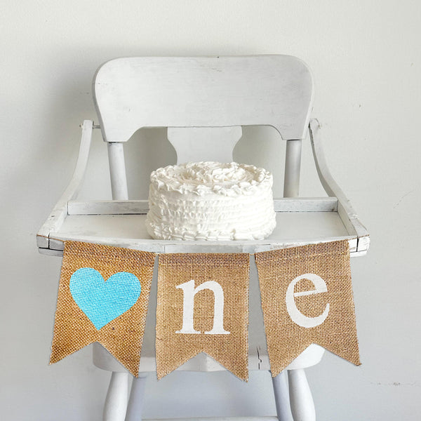 A burlap banner with a blue painted hearts and the letters n and e, making the banner spell one with the heart as the o.