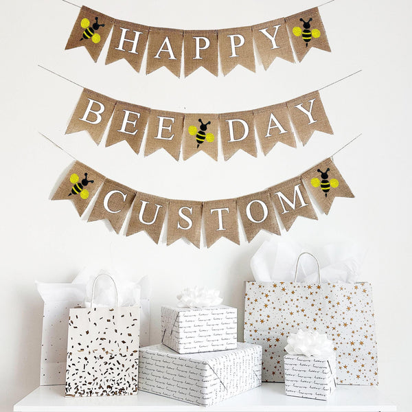 A two tiered burlap banner. The first tier reads HAPPY with a yellow bee icon on either side of the word. The second tier reads BEE DAY with a yellow bee icon between the words.