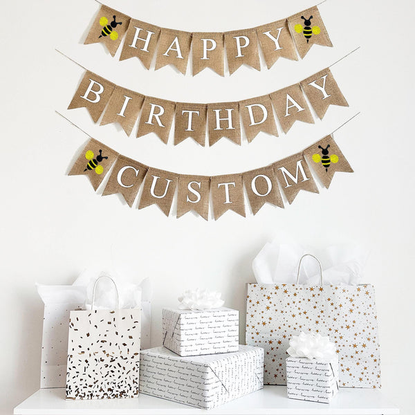 A three tiered burlap banner. The first tier reads HAPPY with a yellow bee icon on either side of the word. The second tier reads Birthday and the third line can be customized to say a name, date or saying with a yellow bee icon between the words.