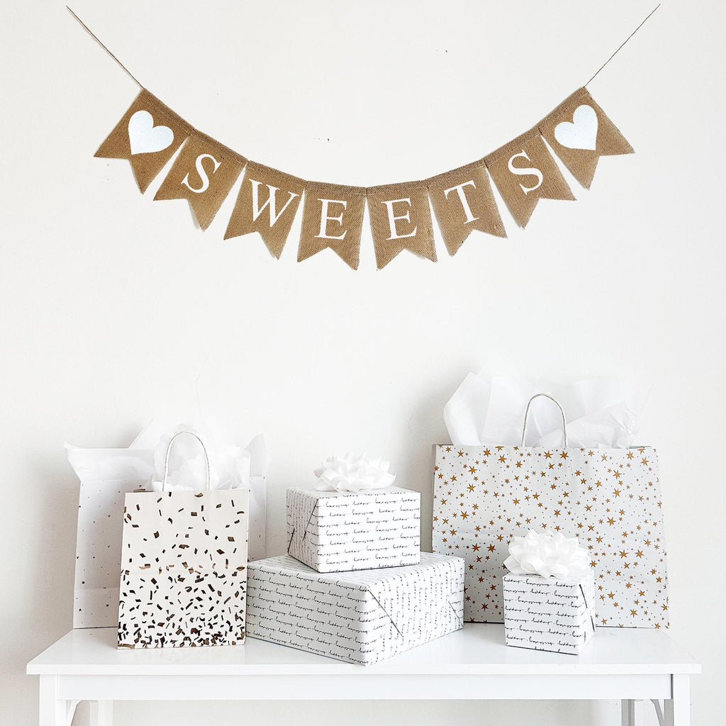 A burlap banner that spells out the word SWEETS in white letters. On each end of the word there is a hand stenciled white heart. Each fishtail burlap flag measures 4.5 x 6.5 inches.