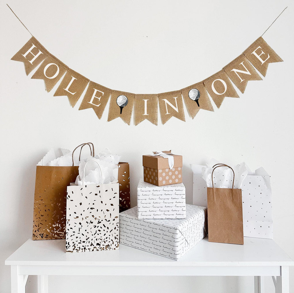 A burlap banner made from 4.5 by 6.5 inch fishtail shaped flags. Each flag has a pocket across the top for stringing. It says HOLE IN ONE in white capital letters and has a hand stenciled image of a white golf ball sitting on a black tee in between each of the words. It is strung on a jute string that is 110 inches in length.