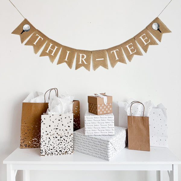 A burlap banner made from 4.5 by 6.5 inch fishtail shaped flags. Each flag has a pocket across the top for stringing. It says THIR-TEE in white capital letters and has a hand stenciled image of a white golf ball sitting on a black tee on each end. It is strung on a jute string that is 110 inches in length.