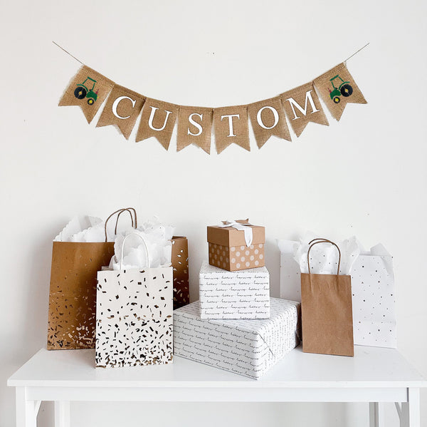 A burlap banner made from 4.5 by 6.5 inch fishtail shaped flags. Each flag has a pocket across the top for stringing. Add a custom name or word of your choice in white capital letters. There is an image of a green tractor on each end of the word. It is strung on a 110 inch jute string.