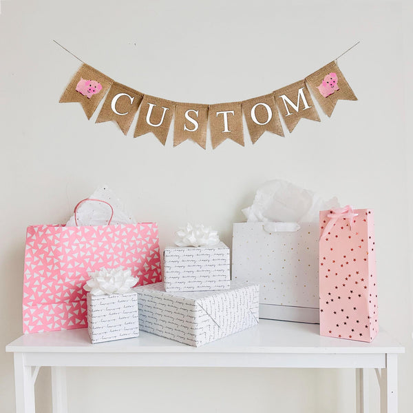 A burlap banner made from 4.5 by 6.5 inch fishtail shaped flags. Each flag has a pocket across the top for stringing. Add a custom name or word of your choice in white capital letters. There is a hand stenciled image of a pink pig on each end of the word. It is strung on a 110 inch jute string.