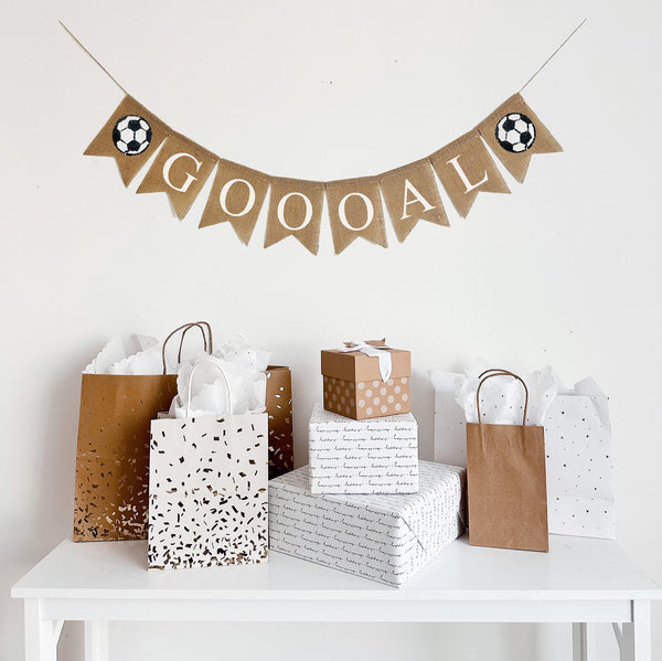 A burlap banner that says &quot;GOOOAL&quot; in white capital lettering with black and white soccer ball images on either side of the word.