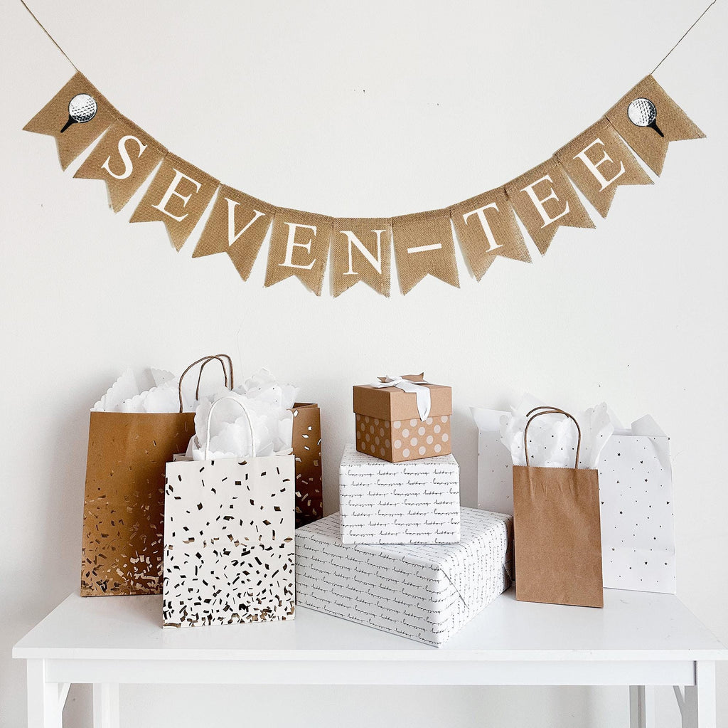 A burlap banner made from 4.5 by 6.5 inch fishtail shaped flags. Each flag has a pocket across the top for stringing. It says SEVEN-TEE in white capital letters and has a hand stenciled image of a white golf ball sitting on a black tee on each end. It is strung on a jute string that is 110 inches in length.