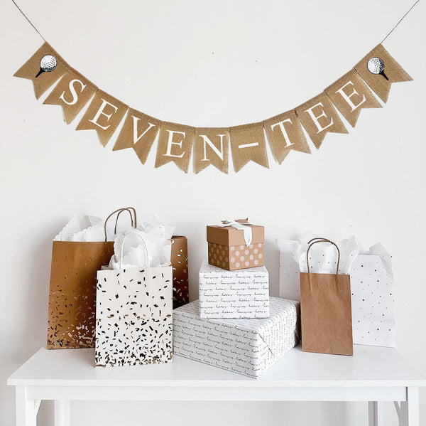A burlap banner made from 4.5 by 6.5 inch fishtail shaped flags. Each flag has a pocket across the top for stringing. It says SEVEN-TEE in white capital letters and has a hand stenciled image of a white golf ball sitting on a black tee on each end. It is strung on a jute string that is 110 inches in length.