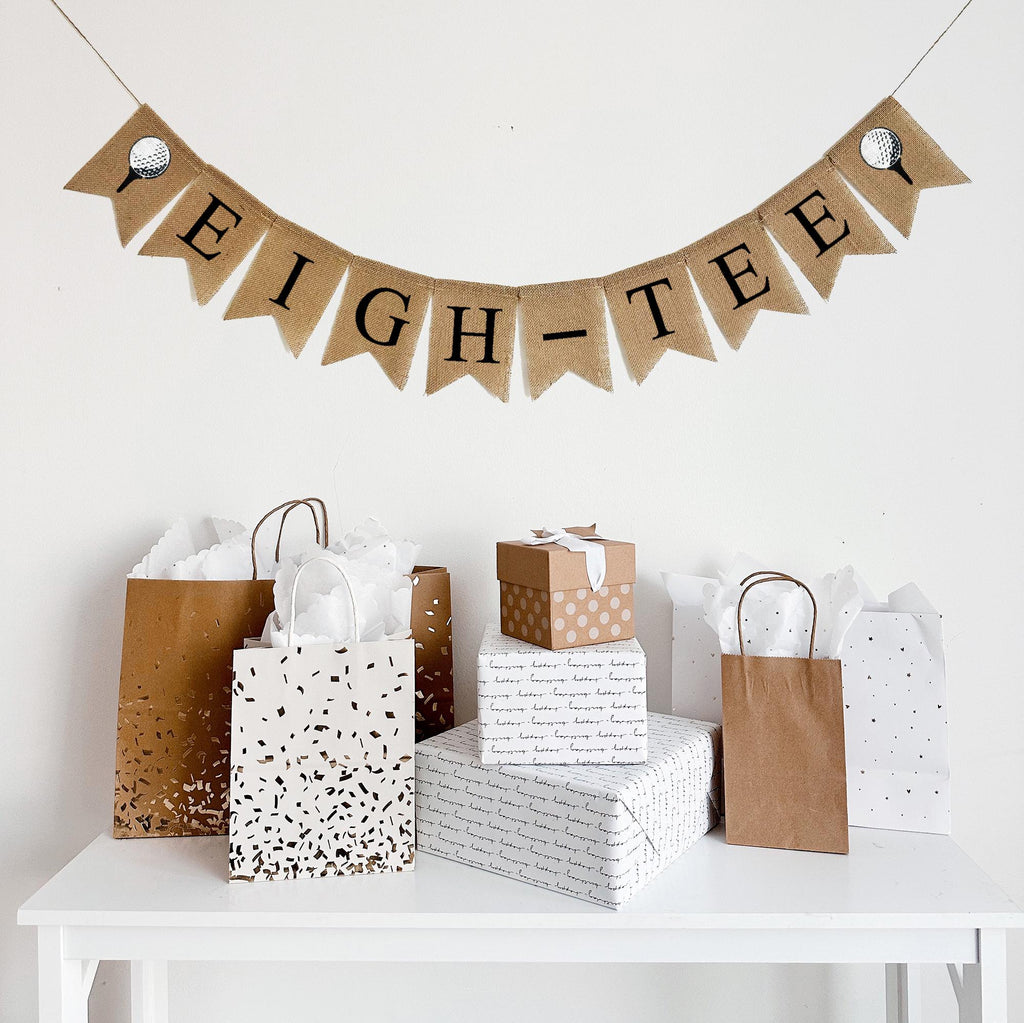 A burlap banner made from 4.5 by 6.5 inch fishtail shaped flags. Each flag has a pocket across the top for stringing. It says EIGH-TEE in black capital letters and has a hand stenciled image of a white golf ball sitting on a black tee on each end. It is strung on a jute string that is 110 inches in length.