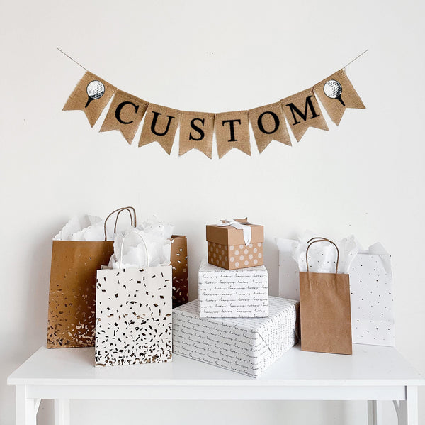 A burlap banner made from 4.5 by 6.5 inch fishtail shaped flags. There are pockets sewn across the top of each flag for stringing. There is an image of a golf ball on a tee on either end. Choose your own custom name or word in black capital letters to personalize your banner. It is strung on a jute string that is 110 inches in length.