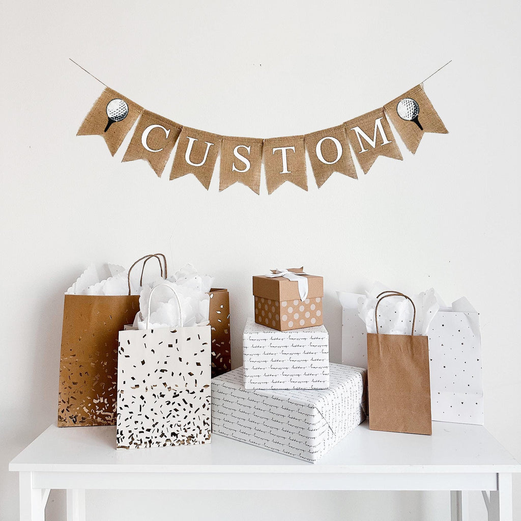 A burlap banner made from 4.5 by 6.5 inch fishtail shaped flags. There are pockets sewn across the top of each flag for stringing. There is an image of a golf ball on a tee on either end. Choose your own custom name or word in white capital letters to personalize your banner. It is strung on a jute string that is 110 inches in length.
