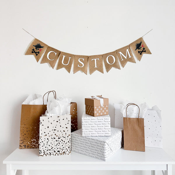 A burlap banner made from 4.5 by 6.5 inch fishtail shaped flags. Each flag has a pocket across the top for stringing. Add a custom name or word of your choice in white capital letters. There is a hand stenciled image of a skull and crossbones wearing a pirate hat on each end of the word. It is strung on a 110 inch jute string.