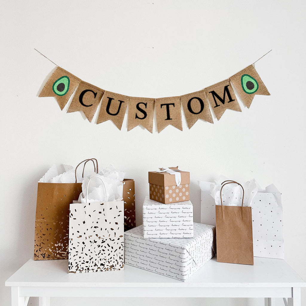 A burlap banner with the word CUSTOM in black lettering between two avocado image flags.