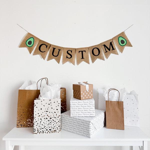 A burlap banner with the word CUSTOM in black lettering between two avocado image flags.