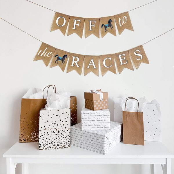 A burlap banner made from 4.5 by 6.5 inch fishtail shaped flags. Each flag has a pocket across the top for stringing. The top string of this banner says OFF in white capital letters and TO in white script one on flag with a hand stenciled image of a horse with a blue saddle in between the words. The bottom string says THE in white script on one flag and RACES  in white capital letters with a hand stenciled horse with brown saddle in between the words. It is strung on two 110 inch jute strings.