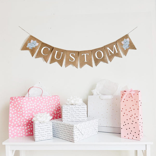 A burlap banner made from 4.5 by 6.5 inch fishtail shaped flags. Each flag has a pocket across the top for stringing. This is a custom banner in white capital letters. Add the name of your choosing to personalize this banner. There is a hand stenciled image of a grey elephant with pink ears on each end of the name. It is strung on a jute string that is 110 inches in length.