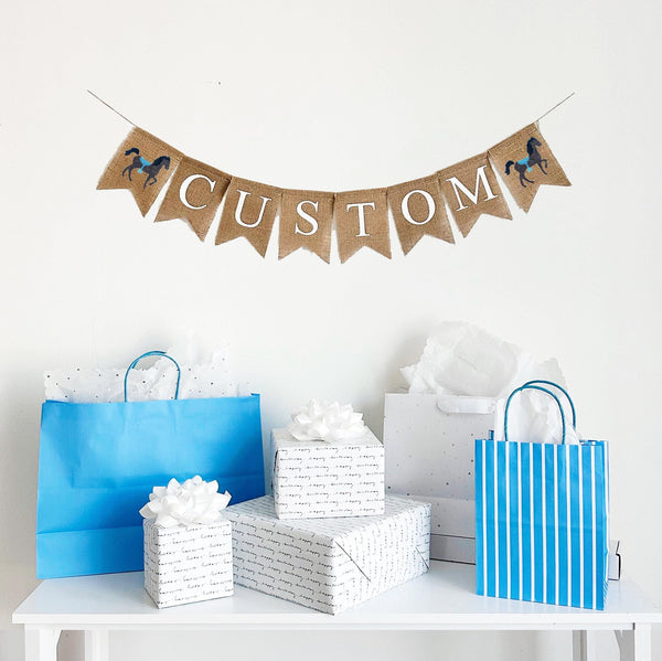 A burlap banner made from 4.5 by 6.5 inch fishtail shaped flags. Each flag has a pocket across the top for stringing. This is a custom banner in white capital letters with a hand stenciled image of a brown horse with a blue saddle on each end. Add a name or word of your choosing to customize the banner. It is strung on a 110 inch jute string.