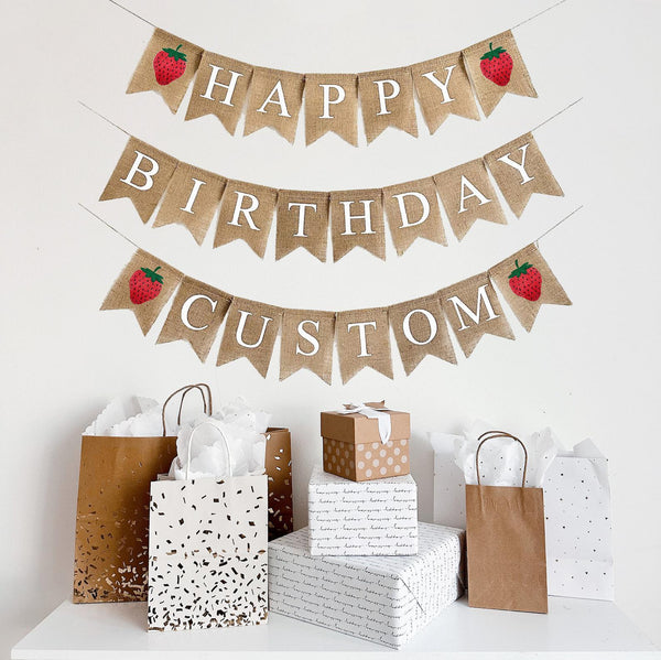A happy birthday burlap banner strung on two or three strings. The happy is at the top and the birthday below.  Hand painted strawberries are at the beginning and end of the word happy.  The strawberries are painted in red with a green stem and black seeds. This banner has the option to add a third line with a custom name or saying with two additional strawberry image flags.