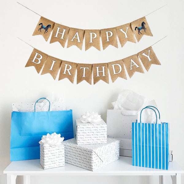 A burlap banner made from 4.5 by 6.5 inch fishtail shaped flags. Each flag has a pocket across the top for stringing. The top line of this banner says HAPPY in white capital letters on the top line with a hand stenciled image of a brown horse with a blue saddle on each end. The bottom line says BIRTHDAY in white capital letters. It is strung on two 110 inch jute strings.
