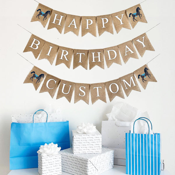 A burlap banner made from 4.5 by 6.5 inch fishtail shaped flags. Each flag has a pocket across the top for stringing. The top line says HAPPY in white capital letters with a hand stenciled image of a brown horse with a blue saddle on each end. The middle line says BIRTHDAY in white capital letters. The bottom line can be customized with a name or word of your choosing in white capital letters and also has a horse image on each end. It is strung on three 110 inch jute strings.