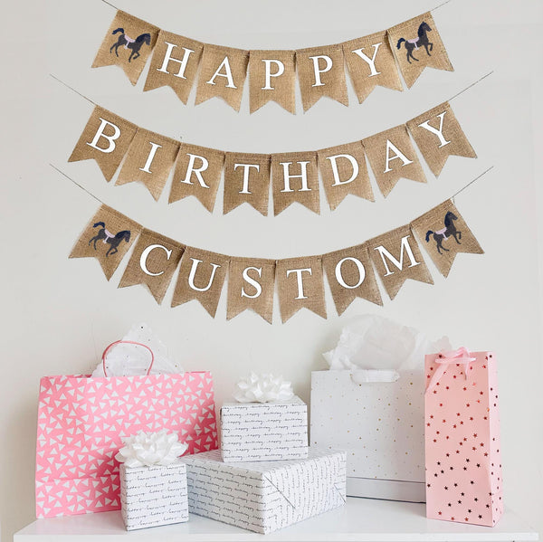 A burlap banner made from 4.5 by 6.5 inch fishtail shaped flags. Each flag has a pocket across the top for stringing. The top line says HAPPY in white capital letters with a hand stenciled image of a brown horse with a pink saddle on each end. The middle line says BIRTHDAY in white capital letters. The bottom line can be customized with a name or word of your choosing in white capital letters and also has a horse image on each end. It is strung on three 110 inch jute strings.
