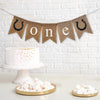 A burlap banner made from 4.5 by 6.5 inch fishtail shaped flags. Each flag has a pocket across the top for stringing. This banner says ONE in white lowercase letters and has a hand stenciled image of a black horseshoe with with nails on each end. It is strung on a 110 inch jute string.