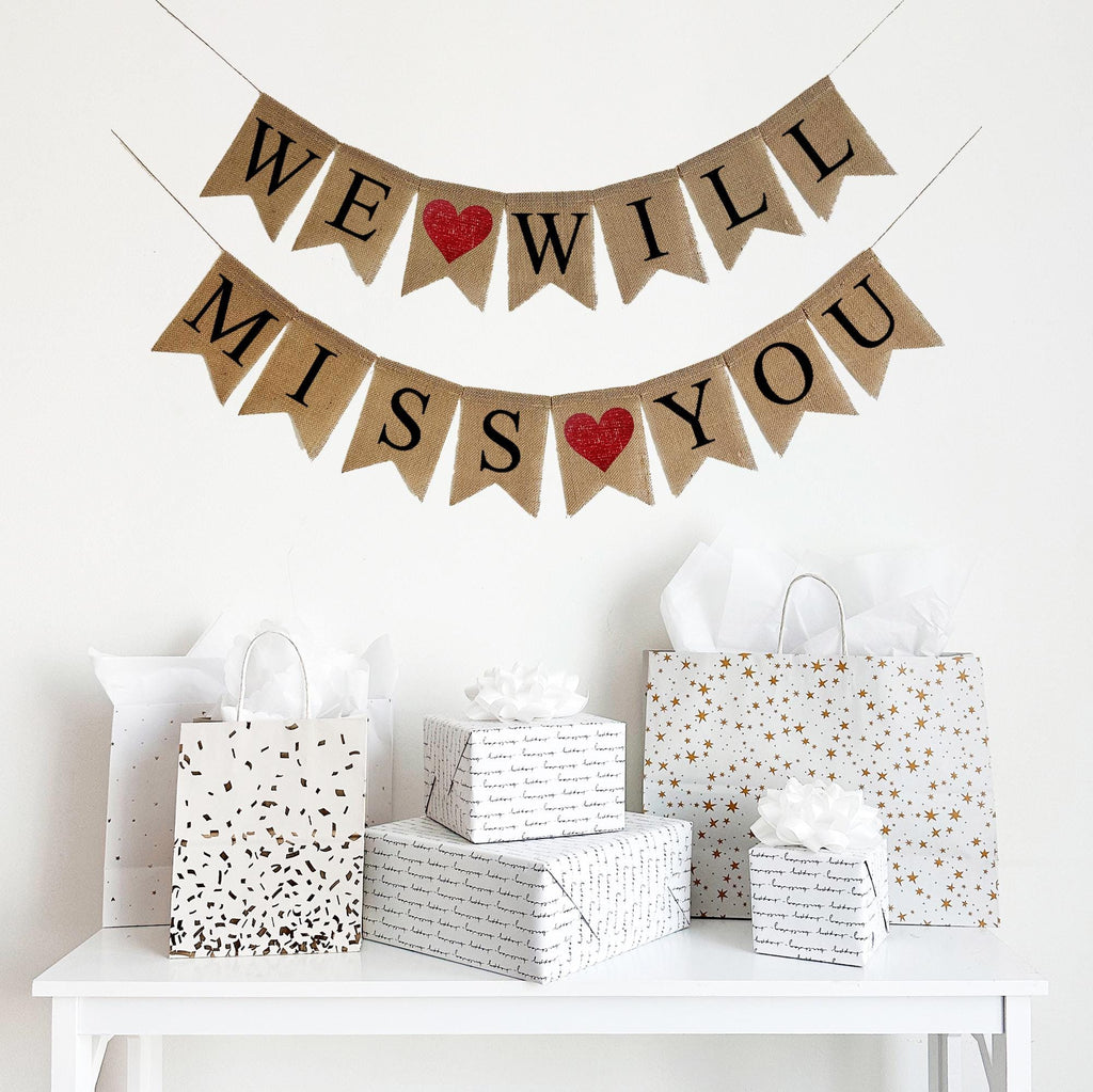 A burlap banner made from 4.5 by 6.5 inch fishtail shaped flags. Each flag has a pocket across the top for stringing. The top line of this banner says WE WILL in black capital letters with a red heart between the words. The bottom line says MISS YOU in black capital letters with a red heart between the words. It is strung on two 110 inch jute strings.