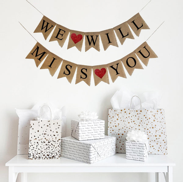 A burlap banner made from 4.5 by 6.5 inch fishtail shaped flags. Each flag has a pocket across the top for stringing. The top line of this banner says WE WILL in black capital letters with a red heart between the words. The bottom line says MISS YOU in black capital letters with a red heart between the words. It is strung on two 110 inch jute strings.