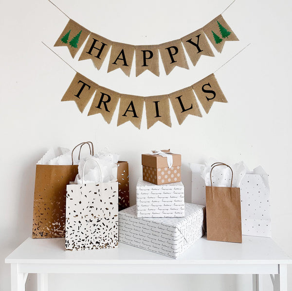A burlap banner made from 4.5 by 6.5 inch fishtail shaped flags. Each flag has a pocket across the top for stringing. The top line of this banner says HAPPY in black capital letters with a hand stenciled image of two green pine trees on each end of the word. The bottom line says TRAILS in black capital letters. It is strung on two 110 inch jute strings.