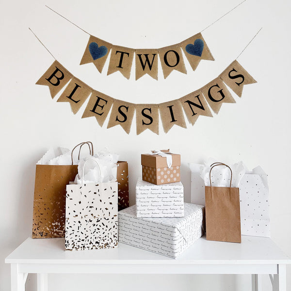 A burlap banner made from 4.5 by 6.5 inch fishtail shaped flags. Each flag has a pocket across the top for stringing. The top line says TWO in black capital letters and has a navy blue heart on each end. The bottom line says BLESSINGS in black capital letters. It is strung on two 110 inch jute strings.