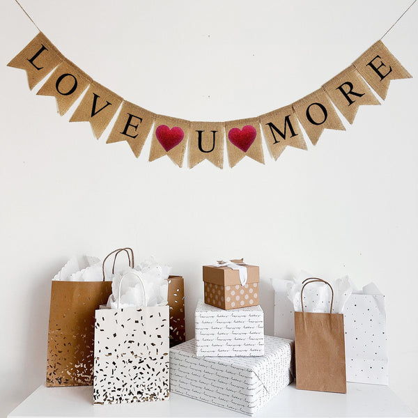 This burlap banner hangs on one line and ready Love U More in black lettering on fishtail burlap flags. Between each word there is a red glitter heart. There are a total of two red glitter hearts, one between Love and U and one between U and More.