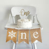 A burlap banner made from 5 by 7 inch fishtail shaped flags. Each flag has a pocket across the top for stringing. This banner says ONE in white lowercase letters and has an image of a glitter snowflake in place of the letter O. It is strung on a 110 inch jute string. It is pictured on a white high chair.