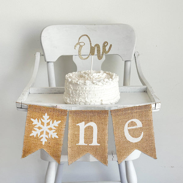 A burlap banner made from 5 by 7 inch fishtail shaped flags. Each flag has a pocket across the top for stringing. This banner says ONE in white lowercase letters and has an image of a glitter snowflake in place of the letter O. It is strung on a 110 inch jute string. It is pictured on a white high chair.