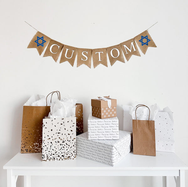 A burlap banner made from 4.5 by 6.5 inch fishtail shaped flags. Each flag has a pocket across the top for stringing. This is a custom banner in white capital letters and has a hand stenciled image of a blue Star of David on each end. Add a name or wording of your choice. It is strung on a 110 inch jute string.