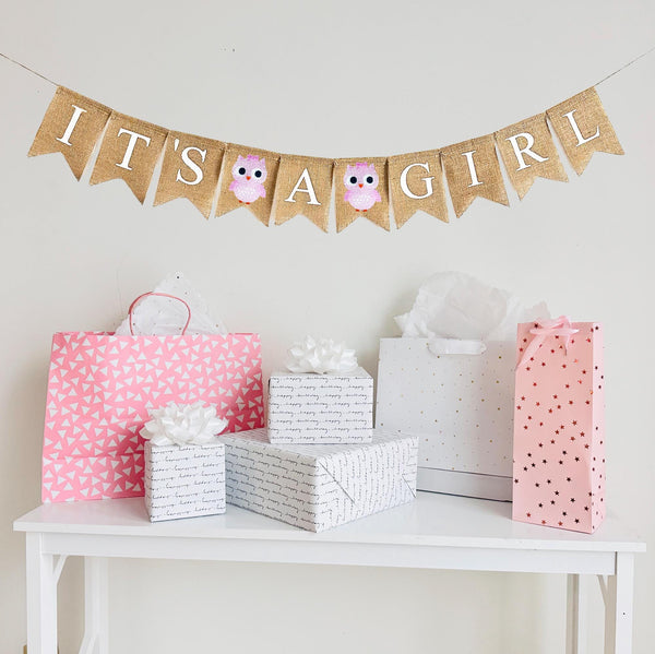 A burlap banner made from 4.5 by 6.5 inch fishtail shaped flags. Each flag has a pocket across the top for stringing. The banner says IT&#39;S A GIRL in white capital letters. There is a hand stenciled image of a pink owl between the words.  It is strung on a jute string that is 110 inches in length.