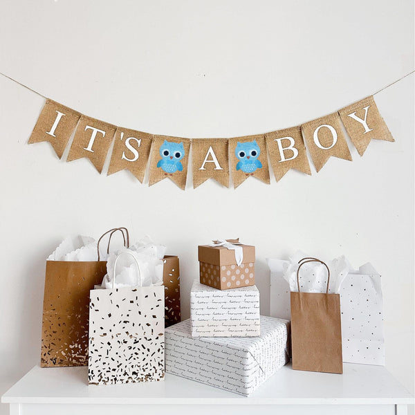 A burlap banner made from 4.5 by 6.5 inch fishtail shaped flags. Each flag has a pocket across the top for stringing. The banner says IT&#39;S A BOY in white capital letters. There is a hand stenciled image of a blue owl between the words.  It is strung on a jute string that is 110 inches in length.