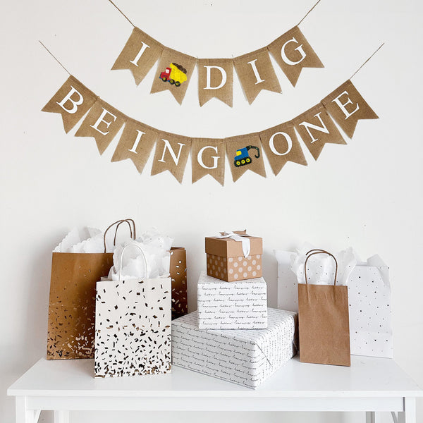 This two tiered banner spells out the words I Dig Being One. Between the words I and Dig there is a red and yellow dump truck image. The second line reads Being One with a blue digger truck in between the words Being and One.