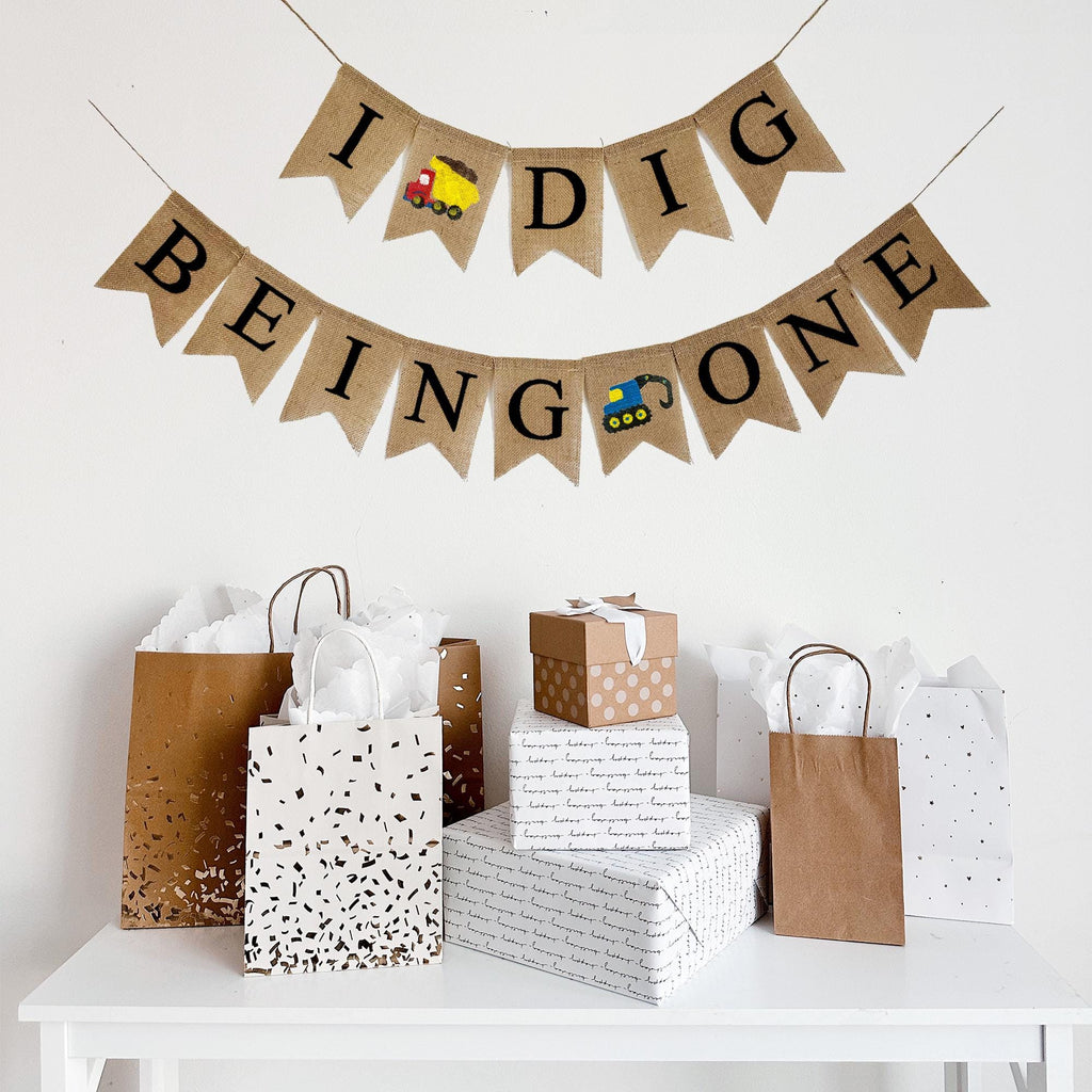 This two tiered banner spells out the words I Dig Being One. Between the words I and Dig there is a red and yellow dump truck image. The second line reads Being One with a blue digger truck in between the words Being and One.