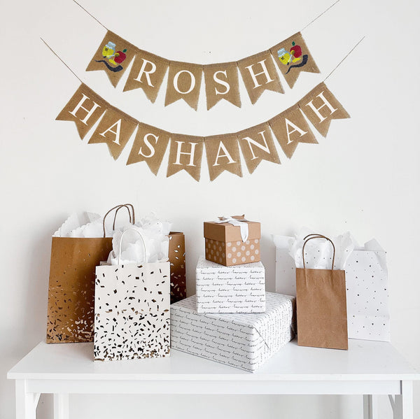 A burlap banner made from 4.5 by 6.5 inch fishtail shaped flags. Each flag has a pocket across the top for stringing. This banner says ROSH on the top line in white capital letters and has a hand stenciled image of a shofar, apple and honey jar on each end of the word. The bottom line says HASHANAH in white capital letters. It is strung on two 110 inch jute strings.