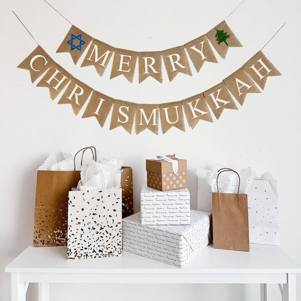 A burlap banner made from 4.5 by 6.5 inch fishtail shaped flags. Each flag has a pocket across the top for stringing. This banner says MERRY in white capital letters and has a hand stenciled image of a blue Star of David on the left end and a green Christmas tree with red decorations on the right side. The bottom line says CHRISMUKKAH in white capital letters. It is strung on two 110 inch jute strings.