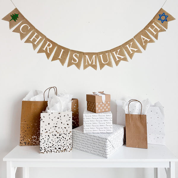 A burlap banner made from 4.5 by 6.5 inch fishtail shaped flags. Each flag has a pocket across the top for stringing. This banner says CHRISMUKKAH in white capital letters and has a hand stenciled image of a green Christmas tree with red decorations on the left side and a blue Star of David on the right side. It is strung on a 110 inch jute string.