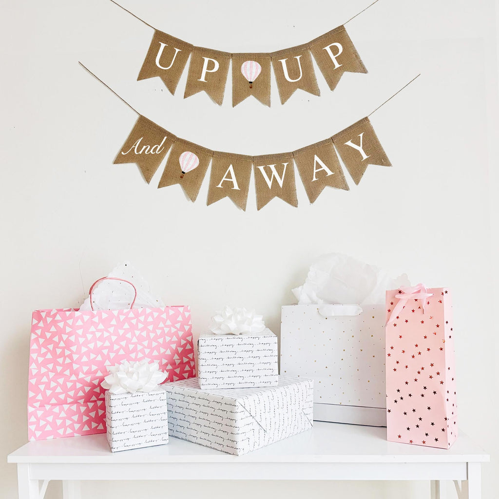 This two tiered Up Up & Away banner add a whimsical touch to your event. The first lay of the banner reads Up Up with a pastel pink striped hot air balloon. The second line starts with the word and stenciled in a script font on one flag followed by another pink hot air balloon and the words Away.
